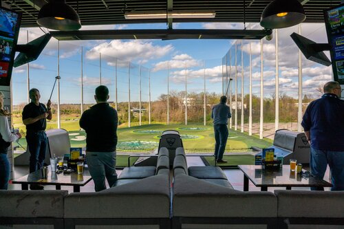 The image depicts an indoor golf entertainment venue, specifically a Topgolf location. The setting is an open-air driving range with individual bays where people can practice their golf swings. The scene is lively, with several individuals engaged in golfing activities. Each person is holding a golf club, preparing to hit golf balls towards a large, open field with multiple targets.

The environment is modern and well-lit, with a combination of natural light streaming in from the large open windows and artificial lighting from overhead fixtures. The sky is partly cloudy, adding a dynamic backdrop to the scene. The range is equipped with tall netting to contain the golf balls, and the targets on the field are circular and color-coded, designed to score points based on accuracy.

In the foreground, there are tables with drinks, suggesting a social and recreational atmosphere. The venue is designed for both entertainment and relaxation, offering a casual setting for friends or family gatherings. The overall mood is energetic and fun, emphasizing leisure and enjoyment.

The presence of multiple screens and modern seating arrangements highlights the venue's blend of sports and social interaction, catering to both avid golfers and casual visitors. The setting is indicative of a popular trend in combining sports with social activities, making it a distinctive and appealing environment for diverse groups.
