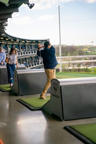 The image depicts a lively scene at a Topgolf venue, a popular entertainment and sports complex known for its driving range games. In the foreground, a man is captured mid-swing, indicating he is actively participating in a golf game. He is dressed in a navy jacket and khaki pants, suggesting a casual, relaxed atmosphere. 

To his left, a woman is preparing to take her turn, holding a golf club and wearing a blue and white outfit. Her stance and focus suggest she is engaged in the activity. The setting features multiple driving bays, each equipped with green turf mats and barriers branded with the Topgolf logo, emphasizing the venue's identity.

The background reveals a multi-level structure with numerous bays, indicating a bustling environment with several participants. The outdoor setting is bright, with natural light illuminating the scene, suggesting a daytime activity. The sky is partly cloudy, adding to the pleasant ambiance.

The overall atmosphere is one of leisure and enjoyment, typical of recreational outings. The presence of modern facilities and the structured layout of the bays highlight the venue's focus on providing a fun and interactive experience for guests.