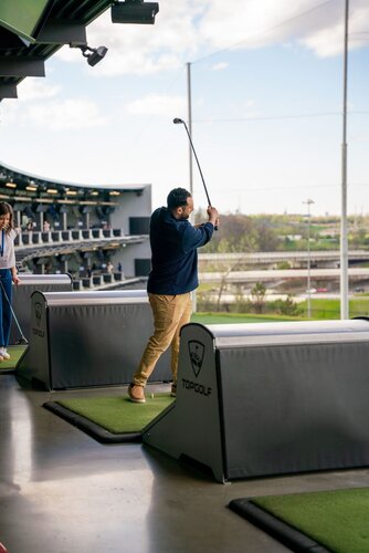 The image depicts a scene at a Topgolf venue, a popular entertainment complex that combines a driving range with a social atmosphere. The setting is modern and open, with multiple levels visible in the background, each equipped with golf bays. The main focus is on a man swinging a golf club, suggesting he is in the middle of a game. He is wearing a navy blue sweater and khaki pants, standing on a green mat designated for players.

The Topgolf logo is prominently displayed on the barriers separating the bays, reinforcing the brand's identity. The background shows a clear sky with some clouds, and the area is well-lit, indicating it might be daytime. The atmosphere is casual and recreational, typical of a leisure activity setting.

The presence of other individuals in the background, possibly preparing to play or watching, adds to the social aspect of the venue. The overall mood is relaxed and enjoyable, capturing the essence of a fun outing with friends or family. The image conveys a sense of motion and activity, characteristic of sports and entertainment environments.