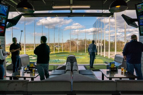 The image depicts an indoor golf entertainment venue, likely a Topgolf location, characterized by its distinctive driving range setup. The scene is set in a spacious, open-air lounge area with modern, comfortable seating, including couches and tables, where patrons can relax and enjoy refreshments. 

In the foreground, several individuals are engaged in golfing activities. One person is poised with a golf club, suggesting they are about to take a swing. Others are observing or preparing for their turn. The patrons are casually dressed, indicating a relaxed, recreational atmosphere.

The backdrop features a large, open driving range with a series of targets and nets, designed for practicing golf shots. The range is surrounded by tall netting to contain the golf balls. The sky is clear with scattered clouds, and the lighting suggests a bright, sunny day, enhancing the vibrant, lively ambiance.

Prominent branding is visible, such as the Topgolf logo on various surfaces, reinforcing the venue's identity. The setting conveys a social and leisurely environment, ideal for gatherings or casual outings, blending sports with entertainment.