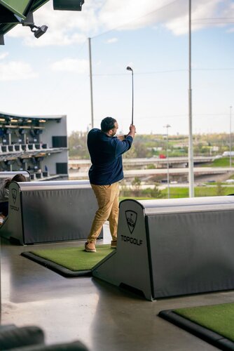 The image depicts a man engaged in a golfing activity at a Topgolf venue. He is positioned on a driving range platform, swinging a golf club. The man is wearing a navy blue jacket and khaki pants, and he appears focused on his swing. The setting is an open-air driving range with multiple levels, typical of Topgolf locations, which are known for combining golf with entertainment and socializing.

The Topgolf logo is visible on the barriers separating the individual bays, reinforcing the venue's branding. The background shows a clear sky with some clouds, and a view of the surrounding area, including a highway and some greenery, suggesting an urban or suburban location.

The lighting is natural, with daylight illuminating the scene, creating a bright and energetic atmosphere. The overall mood is casual and recreational, typical of a leisure activity where individuals or groups gather for fun and relaxation. The image captures the essence of a modern, social golfing experience, appealing to both golf enthusiasts and casual players.