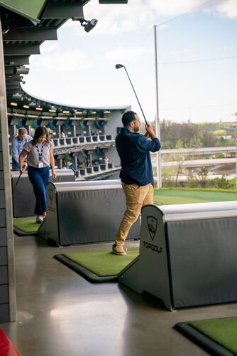 The image depicts a scene at a Topgolf venue, a popular entertainment complex known for its driving range games. In the foreground, a man is captured mid-swing with a golf club, indicating he is actively participating in a golf game. He is dressed casually in a navy shirt and khaki pants. Behind him, a woman and another man are preparing to take their turns, with the woman holding a golf club. 

The setting is an open-air driving range with multiple levels visible in the background, showcasing several other players engaged in similar activities. The area is equipped with golf mats and barriers branded with the Topgolf logo. The lighting is natural, suggesting daytime, with a clear sky visible through the open sides of the venue. 

The atmosphere is lively and recreational, typical of a social sporting event. The venue's modern design and the presence of multiple participants indicate a casual, fun environment where people gather for leisure and entertainment. The image captures the essence of a contemporary social activity, blending sports with a relaxed, communal experience.