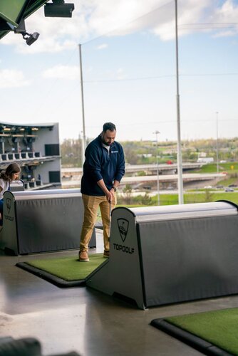 The image depicts a man at a Topgolf venue, a popular entertainment complex that combines a driving range with a lounge-like atmosphere. He is preparing to swing a golf club, standing on a green mat within a designated hitting bay. The Topgolf logo is visible on the partition in front of him, indicating the brand and setting.

The man is dressed casually in a navy blue pullover and khaki pants, focusing on his stance and grip on the club. The setting is open-air, with multiple levels visible, suggesting a multi-tiered structure typical of Topgolf locations. In the background, there are other bays with people engaged in similar activities, and a view of the outdoors with a clear sky and some clouds.

The lighting is natural, suggesting daytime, and the atmosphere is relaxed and recreational, typical of leisure activities. The setting conveys a modern, social environment where people gather for casual golfing and entertainment.
