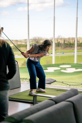 The image depicts a woman engaged in a golfing activity at a driving range. She is positioned on a green mat, holding a golf club mid-swing, suggesting she is about to hit a golf ball. Her attire consists of a light gray top and blue pants, paired with white sneakers, indicating a casual and relaxed setting. The background reveals a well-maintained golf course with neatly trimmed grass and sand bunkers, typical of a driving range environment.

The setting is outdoors, with a clear sky visible, suggesting a pleasant day with good weather. The lighting is natural, contributing to a bright and inviting atmosphere. The presence of tall netting poles in the background indicates safety measures typical of driving ranges to contain golf balls.

The overall mood of the image is leisurely and recreational, capturing a moment of enjoyment and focus. The scene conveys a sense of casual sporting activity, possibly during a social or leisure event. The environment and activity suggest a modern, recreational facility designed for both practice and entertainment.