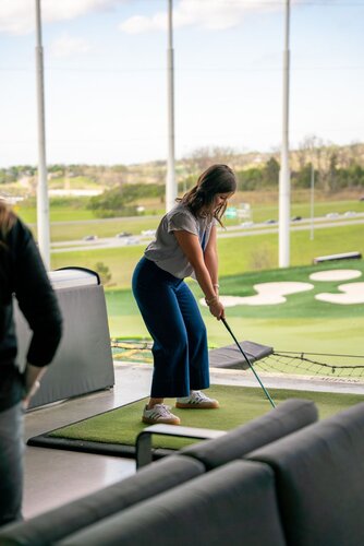 The image depicts a woman preparing to swing a golf club at an indoor driving range. She is positioned on a green mat, holding a golf club, and appears focused on her stance. The setting is a modern golf facility with an open view of the outdoor range, featuring well-maintained grass and several targets or markers in the distance. The sky is clear, suggesting a bright day, which adds a fresh and lively atmosphere to the scene.

The woman is dressed casually in a gray top and blue pants, with white sneakers, embodying a relaxed yet sporty look. Her posture indicates concentration and readiness, typical of someone about to take a golf swing. The background includes a view of the expansive golf range, with visible netting and poles that define the boundaries of the facility.

The environment suggests a leisurely activity, possibly recreational or practice-oriented, rather than a competitive event. The presence of couches and a social setting implies a space designed for both playing and relaxation, common in entertainment venues like Topgolf, which combines golfing with a social experience.

Overall, the image conveys a theme of leisure and sport, capturing a moment of focus and enjoyment in a contemporary recreational setting.