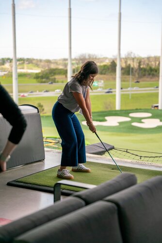 The image depicts a young woman preparing to swing a golf club at an indoor driving range. She stands on a green mat, holding the club with both hands, poised to hit a golf ball. The setting is modern and open, with large windows revealing an outdoor view of a golf course. The course features several sand traps and a well-maintained green, with a highway visible in the background, indicating a suburban or urban location.

The woman is dressed casually in a gray shirt and blue pants, with white sneakers, suggesting a relaxed, recreational activity. The lighting is natural, with daylight streaming in, creating a bright and inviting atmosphere. The presence of couches and a barrier netting around the area indicates a social, leisure-focused environment, possibly a venue like Topgolf, known for combining golf with entertainment.

The image conveys a theme of casual recreation and social interaction, capturing a moment of concentration and anticipation as the woman prepares to play. The setting suggests a blend of sport and leisure, appealing to both golf enthusiasts and those seeking a fun outing.