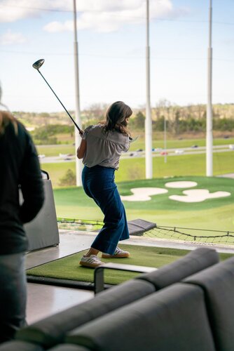 The image depicts a woman engaged in a golfing activity at a driving range. She is captured mid-swing, holding a golf club, with her body slightly turned away from the camera. The setting is an outdoor driving range, characterized by a series of targets and sand traps visible in the distance. The area is surrounded by a netted barrier for safety.

The woman is dressed casually in a gray blouse and blue jeans, paired with sneakers, suggesting a relaxed, recreational atmosphere. The lighting is natural, indicating a daytime setting with clear skies and some clouds visible, contributing to a bright and open ambiance.

In the background, a stretch of green grass and a few trees can be seen, along with a road where a few cars are driving, adding a sense of motion and life to the scene. The presence of couches and a railing in the foreground suggests a comfortable, social environment typical of modern driving ranges that cater to both serious golfers and casual visitors.

The overall mood of the image is leisurely and active, capturing a moment of enjoyment and focus in a recreational sports setting.