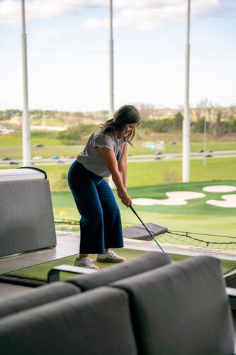 The image depicts a woman engaged in a golfing activity at an indoor driving range, likely a facility like Topgolf. She is positioned on a platform, preparing to swing a golf club. The setting includes a green artificial turf mat and a safety net in front of her, which is characteristic of driving ranges designed for practice and entertainment.

The background shows an expansive view of a golf course with several targets on the ground, indicating areas for players to aim their shots. Beyond the course, there is a landscape of grassy fields and a road with visible traffic, suggesting the facility is located in a suburban or semi-urban area.

The woman is dressed casually in a light gray top and dark blue pants, with white sneakers, embodying a relaxed and recreational atmosphere. The lighting is natural, suggesting daytime, with a clear sky visible through large open windows or bays, enhancing the open and airy feel of the environment.

The overall mood of the image is leisurely and recreational, emphasizing enjoyment and relaxation in a social or family-friendly setting. The image captures the essence of a modern, interactive golfing experience, blending sport with entertainment.