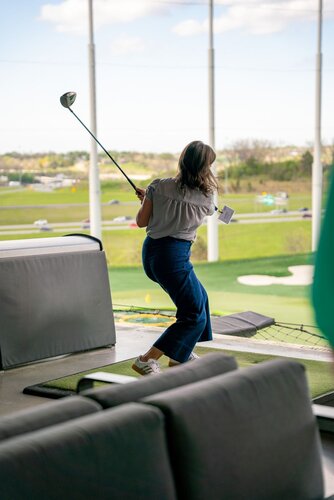 The image depicts a woman swinging a golf club at a driving range. She is positioned on a mat, mid-swing, with her back slightly turned towards the camera. The setting is outdoors, with a view of a green expanse and a highway in the background, indicating a suburban or semi-urban location. The driving range features several levels, and she is on an upper level, as indicated by the netting and barriers.

The woman is dressed casually in a light gray blouse and dark blue pants, paired with white sneakers. Her posture suggests she is focused and engaged in the activity. The golf club is a driver, typically used for long-distance shots.

The lighting is natural, suggesting daytime, with a clear sky and a few clouds visible. The overall atmosphere is relaxed and recreational, typical of a leisure activity setting. The image captures a moment of action and concentration, with the woman likely aiming to hit the golf ball towards the targets on the range.

This scene is reminiscent of entertainment venues like Topgolf, which combine golfing with a social atmosphere, though no specific branding is visible to confirm this. The image conveys a sense of leisure and enjoyment, characteristic of a casual outing or social gathering.