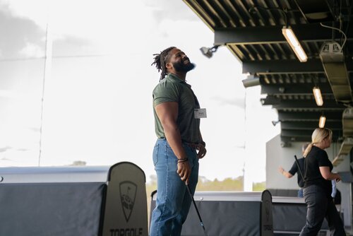 The image depicts a man standing in an indoor golf driving range. He is holding a golf club, suggesting he is about to take a swing or has just completed one. The man is wearing a green polo shirt and blue jeans, and he has a name tag on his chest. His expression appears focused or contemplative, as he looks upward. 

The setting is a modern driving range with overhead lighting and a structured ceiling, creating an industrial yet recreational atmosphere. To the right, another person is visible, possibly preparing to swing. The environment is well-lit, with natural light coming in from the open side of the range, highlighting the contrast between the indoor and outdoor spaces.

The driving range bays are equipped with barriers that have the "Topgolf" logo, indicating the specific venue. The overall mood of the image is relaxed and recreational, typical of a leisure activity like golfing. The scene conveys a sense of focus and enjoyment, characteristic of a casual day spent at a sports facility.