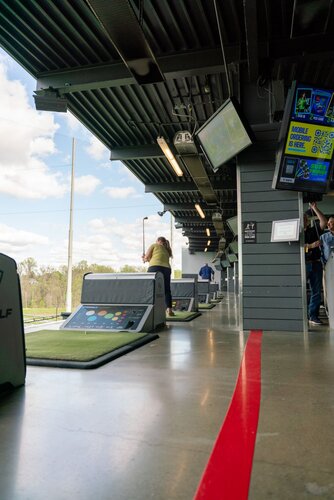 The image depicts an indoor golf driving range, specifically a facility similar to Topgolf. The setting features several hitting bays lined up, each equipped with golf mats and seating areas. A prominent red line runs along the polished concrete floor, guiding the layout of the bays. 

In the foreground, a person is swinging a golf club, indicating active participation in the sport. The individual is positioned in a bay, which includes a digital screen displaying scores and game options, adding a technological aspect to the golfing experience. Above the bays, multiple flat-screen monitors are visible, likely showing game stats or entertainment content, enhancing the interactive atmosphere.

The environment is well-lit with natural light streaming in from the open side, where the driving range extends outdoors. The sky is partly cloudy, suggesting a pleasant day. The overall color scheme includes neutral tones with accents of green from the golf mats and the vibrant red line on the floor.

The setting conveys a modern, recreational atmosphere, blending sports with social interaction. This type of venue is popular for both casual and competitive play, often used for social gatherings or corporate events. The image captures the essence of leisure and entertainment in a contemporary sports facility.