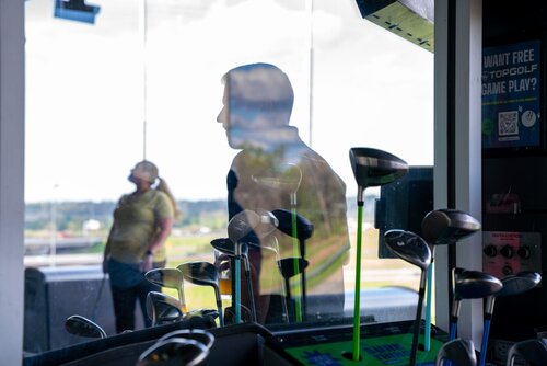 The image depicts an indoor golf facility, likely a Topgolf venue, as suggested by the signage offering free game play. The setting is a modern, recreational environment focused on golf. In the foreground, a variety of golf clubs are prominently displayed, their metallic surfaces reflecting light, adding a dynamic visual element. The clubs are positioned upright, indicating a rental or selection area for players.

In the background, through a large glass window, two individuals are visible. One person is standing in profile, their silhouette partially obscured by reflections on the glass. The other person, further in the background, appears to be looking upwards, possibly observing something outside the frame or enjoying the outdoor view. The setting beyond the glass suggests an open-air driving range, typical of Topgolf locations, with expansive views of the landscape.

The lighting is bright and natural, with sunlight streaming in, creating a contrast between the indoor and outdoor areas. The overall atmosphere is relaxed and recreational, typical of a leisure activity setting. The presence of promotional signage and QR codes hints at a modern, tech-friendly environment, encouraging interaction and engagement.

The image captures a moment of leisure and casual enjoyment, with a focus on the sport of golf in a contemporary, social setting.