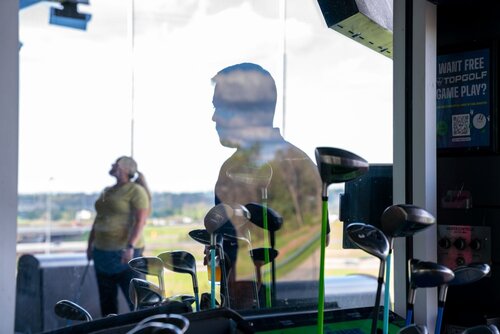 The image depicts an indoor golf setting, likely at a driving range or entertainment venue like Topgolf. In the foreground, there is a rack of golf clubs with various drivers prominently displayed. The clubs are shiny and reflect the ambient light, suggesting a well-lit environment.

In the background, a person is visible through a large window, standing outside in a golf stance, possibly preparing to swing. The silhouette of another individual is reflected in the glass, adding depth to the scene. The setting outside appears to be a golf range, with green fields extending into the distance.

The interior includes a digital screen or poster on the right side, advertising a promotion for free gameplay, indicating a commercial or recreational context. The overall atmosphere is casual and recreational, typical of a leisure activity setting.

The lighting is bright, with natural light streaming in through the windows, creating a contrast between the indoor and outdoor areas. The image conveys a relaxed and engaging environment, inviting for social gatherings or casual sports activities.