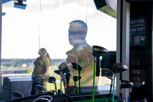 The image depicts an indoor golf setting, likely at a driving range or entertainment venue such as Topgolf. In the foreground, several golf clubs are prominently displayed, with their heads visible, suggesting a focus on golfing activities. The clubs are placed in a rack, ready for use.

In the background, a glass window reflects the silhouette of a person, creating an intriguing visual effect. This reflection adds depth and complexity to the image, as the person is not directly visible but is a key element of the composition. Another individual is seen in the background, standing near the window, which overlooks an outdoor area, possibly a golf course or driving range.

The lighting is bright, with natural light streaming in through the window, casting soft shadows and creating a clear, vibrant atmosphere. The predominant colors are neutral, with hints of green from the golf equipment and the outdoor scenery.

On the right side of the image, there is a sign with text that partially reads "WANT FREE TOPGOLF GAME PLAY?" along with a QR code, indicating a promotional offer or advertisement. This suggests a casual, recreational environment where visitors can enjoy golfing activities and promotions.

The overall atmosphere is relaxed and recreational, typical of a leisure venue where people gather to enjoy sports and social activities. The setting and elements convey a modern, interactive experience associated with entertainment venues like Topgolf, which blend sports with socializing.