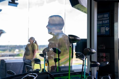 The image depicts an indoor golf entertainment venue, likely a Topgolf location, characterized by its distinctive driving range setup. In the foreground, there is a rack of golf clubs, prominently featuring drivers with metallic heads reflecting the ambient light. The clubs are neatly arranged, indicating an organized and professional setting.

In the background, through large glass windows, two individuals are visible. One person, silhouetted against the bright outdoor light, appears to be engaged in conversation or observing the activity. The other individual, further back, is standing near the edge of the driving range, possibly preparing to take a swing. The outdoor setting beyond the glass shows a clear sky and expansive view, typical of a recreational sports environment.

On the right side of the image, a sign offers "FREE TOPGOLF GAME PLAY," accompanied by a QR code and additional text, suggesting promotional content. The sign's presence indicates a commercial and leisure-oriented atmosphere, inviting participation and engagement.

The lighting is a mix of natural daylight streaming through the windows and artificial lighting inside, creating a contrast between the bright outdoor view and the dimmer interior. The overall mood is casual and recreational, emphasizing social interaction and leisure activities.

The setting is modern and vibrant, with a focus on entertainment and sports, capturing the essence of a popular contemporary pastime.
