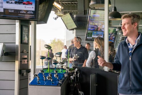 The image depicts a lively scene at a modern golf entertainment venue, likely a driving range. In the foreground, a display of golf clubs is prominently featured, suggesting the setting is designed for recreational golfing. Several individuals are present, engaging in social and leisure activities. One person in the foreground is smiling and holding a golf club, indicating participation in the activity.

Multiple screens are visible, displaying various content, including what appears to be sports-related information and advertisements, enhancing the atmosphere of a sports-centric environment. The lighting is bright and natural, suggesting it is daytime, with large windows allowing ample light into the space.

The attire of the individuals is casual, aligning with the relaxed and recreational nature of the venue. The overall mood is upbeat and social, with people interacting and enjoying the activity. The setting suggests a blend of sports and socializing, typical of entertainment complexes that combine leisure activities with dining and social spaces.