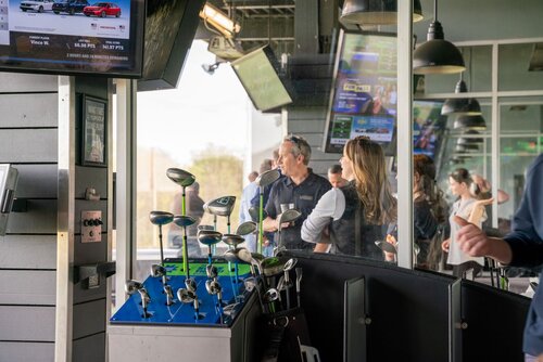The image depicts an indoor golf entertainment venue, likely a driving range such as Topgolf. The setting is modern and lively, with a focus on leisure and social interaction. In the foreground, there is a display of various golf clubs, neatly arranged and ready for use. The clubs are housed in a sleek, metallic stand, suggesting a high-tech and organized environment.

Several individuals are gathered around, engaged in conversation and enjoying the atmosphere. They are casually dressed, indicating a relaxed, recreational setting. The presence of multiple screens displaying sports content and scores adds to the dynamic and interactive ambiance, enhancing the experience for visitors.

The lighting is bright and inviting, with natural light streaming in from large windows, creating a warm and open atmosphere. The predominant colors are neutral tones with splashes of green from the golf equipment, contributing to a fresh and sporty vibe.

Overall, the image conveys a sense of fun and camaraderie, typical of a social sports venue where people gather to enjoy golf in a casual, entertaining environment.