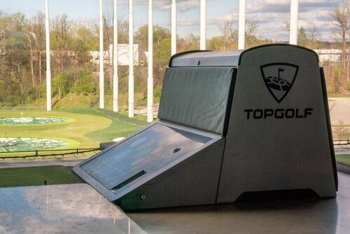 The image depicts a scene from a Topgolf venue, a popular entertainment complex known for its driving range games. The setting is an outdoor area with a view of multiple golf targets on a well-maintained green field. The targets are circular with distinct patterns, likely used for scoring in the game.

In the foreground, there is a prominent Topgolf-branded console, which is used for tracking scores and managing gameplay. The console is sleek and modern, featuring the Topgolf logo, which includes a stylized golf ball and tee. The design is minimalistic, with a gray and black color scheme.

The background shows tall netting surrounding the range, ensuring safety and containment of golf balls. The surrounding environment includes lush greenery and trees, indicating a spring or summer season. The sky is partly cloudy, with sunlight casting a natural, bright illumination over the scene, creating a lively and inviting atmosphere.

This image captures the essence of a recreational activity that combines sports with social interaction, appealing to both golf enthusiasts and casual visitors. The setting is indicative of a leisure activity, likely appealing to groups of friends or families looking for entertainment and relaxation.