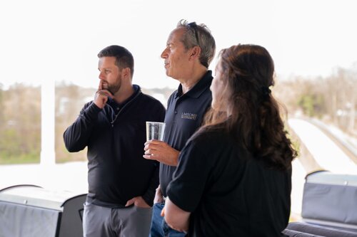 The image depicts three individuals engaged in a casual conversation, possibly at an outdoor or semi-outdoor event. The setting appears to be a covered area with natural light illuminating the scene, suggesting a daytime gathering. The background is slightly blurred, with hints of trees and an open sky, indicating an outdoor environment.

The individuals are dressed in casual attire, with two of them wearing dark polo shirts. One person is holding a clear glass, possibly containing a beverage, which suggests a relaxed, social atmosphere. The man in the center has a thoughtful expression, while the others appear attentive, contributing to a sense of engagement and interaction.

The predominant colors in the image are neutral tones, with the individuals' clothing contrasting against the lighter background. The lighting is soft and natural, enhancing the casual and informal mood of the scene.

The setting and attire suggest a professional yet relaxed event, such as a company outing or a casual business meeting. The overall atmosphere conveys a sense of camaraderie and open communication among the participants.
