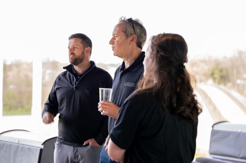 The image depicts three individuals engaged in conversation on what appears to be an outdoor balcony or terrace. The setting is bright, with natural daylight illuminating the scene, suggesting it might be daytime. The background shows a blurred view of a landscape with trees and a road, indicating an elevated vantage point.

The individuals are casually dressed, with two men and one woman. The man in the center is holding a clear glass, possibly containing a beverage, and is wearing a dark polo shirt with a logo that reads "LARSON." The other man, on the left, is wearing a dark jacket, while the woman on the right has her hair tied back and is also wearing a dark top.

The atmosphere appears relaxed and informal, suggesting a casual gathering or event. The individuals seem engaged in a light-hearted conversation, contributing to a friendly and open ambiance. The setting could be part of a social or corporate event, possibly involving networking or leisure activities. The image captures a moment of interaction, highlighting the social aspect of the gathering.
