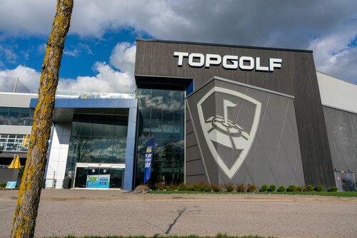The image depicts the exterior of a Topgolf entertainment venue. The building is modern and expansive, featuring a large, prominent logo of a stylized golf ball with a flag, set against a shield-like background. The word "TOPGOLF" is displayed in bold, white letters on the upper section of the building, making it easily identifiable.

The architecture is sleek, with a combination of dark and light gray panels, creating a contemporary and inviting appearance. The entrance is glass-fronted, allowing a glimpse into the interior. There are signs near the entrance, one of which appears to promote an event or offer, although the specific text is not clear.

The setting is an outdoor environment with a clear blue sky and scattered clouds, suggesting a pleasant day. A tree with a textured trunk is visible in the foreground, adding a natural element to the scene. The overall atmosphere is vibrant and energetic, typical of a recreational venue designed for social gatherings and entertainment.

Topgolf is known for its unique blend of golf, dining, and entertainment, often attracting groups for social events or casual outings. The image captures the essence of a leisure destination, combining sports and social interaction in a modern setting.