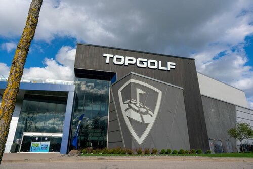 The image depicts the exterior of a Topgolf facility, characterized by its modern architectural design. The building features a large, prominent sign with the text "TOPGOLF" in bold white letters against a dark facade. Below the text is a large emblem resembling a shield with a stylized golf tee and ball, reinforcing the brand identity.

The structure is a mix of glass and metal, with reflective windows on the left side, allowing glimpses of the interior. The sky is partly cloudy, casting a natural light that enhances the building's sleek appearance. A small tree is visible in the foreground, adding a touch of greenery to the scene.

The setting suggests a recreational environment, likely a location where people gather for social and entertainment purposes centered around golf. The atmosphere is inviting and contemporary, indicative of a leisure venue designed for both casual and enthusiastic golfers. The image conveys a sense of modernity and fun, appealing to a wide audience looking for entertainment and social interaction.
