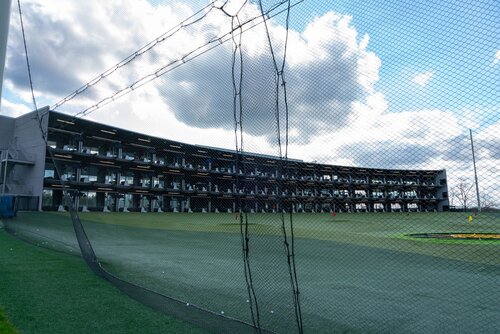 The image depicts a multi-level driving range, likely a Topgolf facility, characterized by its distinctive architectural design. The structure features several tiers of hitting bays, each equipped with seating and golfing equipment. The bays are open to a large, expansive field with clearly marked targets, suggesting a recreational golfing environment.

The setting is outdoors, under a partly cloudy sky, with sunlight casting a natural light across the scene. The driving range is enclosed by a high netting, ensuring safety and containment of golf balls. The grass on the field is well-maintained, contributing to a professional and inviting atmosphere.

The overall mood is relaxed and recreational, typical of leisure sports facilities. The presence of multiple levels and bays indicates a focus on accommodating numerous visitors, suggesting a popular venue for social gatherings and entertainment. The modern design and setup hint at a contemporary, family-friendly environment where individuals or groups can enjoy golfing activities.