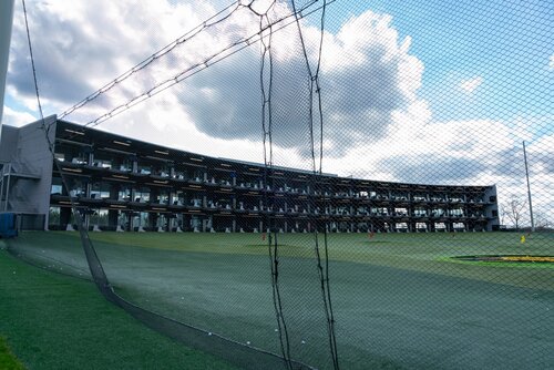 The image depicts a multi-level driving range, likely part of a golf entertainment complex such as Topgolf. The structure features several tiers of hitting bays, each equipped with seating and golf equipment. The building's modern design includes large glass windows and open-air sections for each bay, allowing players to hit golf balls onto a large, open field.

The field is covered with artificial turf and marked with various targets, likely used for scoring in games. A protective netting surrounds the perimeter, ensuring safety and containment of golf balls. The sky is partly cloudy, with a mix of blue and white, suggesting a clear day with good visibility.

The atmosphere is recreational and lively, suggesting a social setting where groups can enjoy both golf and leisure. The design and setup indicate a focus on entertainment and casual play rather than traditional golfing, appealing to a wide range of participants, from novices to experienced players. The overall setting is vibrant and inviting, designed for both daytime and evening activities.