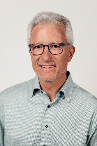 The image is a portrait of an older man with short, gray hair and a neatly trimmed beard. He is wearing blue-framed glasses and a light gray, checkered button-up shirt. The background is a plain, neutral white, which draws focus to the subject. The lighting is soft and even, highlighting the man's facial features without harsh shadows. His expression is friendly and approachable, suggesting a professional or casual setting. The overall atmosphere is calm and composed, typical of a professional headshot used for business or personal branding purposes. The image does not contain any text, symbols, or additional objects, focusing solely on the individual.