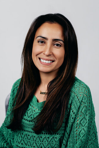 The image features a woman with long, dark hair, smiling warmly at the camera. She is wearing a green, intricately patterned knit sweater, which adds texture and interest to the composition. The background is a plain, light gray, which helps to emphasize the subject without distraction. The lighting is soft and even, highlighting her features gently and creating a welcoming, approachable atmosphere. The overall mood of the image is friendly and professional, suggesting it might be used for a personal or professional profile. The simplicity and clarity of the setting focus attention on the subject, making it suitable for contexts such as corporate headshots or social media profiles.
