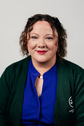 The image features a person with curly brown hair, wearing a bright blue blouse and a dark green cardigan. The cardigan has a small emblem on the left side, which includes a circular design with a star and text that is partially visible. The person is smiling softly, conveying a friendly and approachable demeanor. The background is plain and light-colored, which keeps the focus on the individual. The lighting is even and soft, highlighting the person’s facial features without harsh shadows. The overall atmosphere of the image is professional yet warm, suggesting it might be a portrait for a formal or semi-formal context, such as a professional profile or an organizational representation.