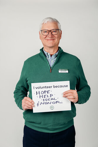 The image features an older man with white hair and glasses, standing against a plain, light-colored background. He is wearing a green zip-up sweater over a checkered shirt, and he has a name tag clipped to his sweater. The man is smiling warmly and holding a sign in front of him that reads, "I volunteer because HOPE, HELP, HEAL, & HONOR." The text on the sign is handwritten in blue ink, emphasizing the personal nature of the message.

The overall atmosphere of the image is positive and uplifting, conveying themes of community service and personal commitment. The man's expression and the words on the sign suggest a sense of dedication and altruism. The setting is neutral, likely intended to keep the focus on the individual and his message.

The image appears to be part of a campaign or initiative promoting volunteerism, possibly for a nonprofit organization or community service group. The simplicity of the background and the clarity of the message contribute to a straightforward and impactful presentation.
