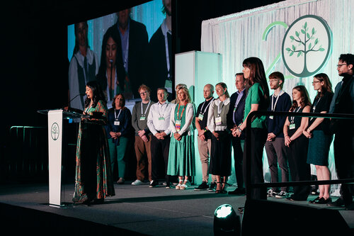 The image depicts a formal event, likely a conference or anniversary celebration, set on a stage with a group of people standing in a line. The backdrop features a large logo with a stylized tree and the number "20," suggesting a 20th anniversary or milestone event. The stage is well-lit with a combination of white and green lighting, creating a professional and celebratory atmosphere.

A woman stands at a podium on the left side of the image, addressing the audience. The podium displays the same tree logo, reinforcing the event's branding. The individuals on stage are dressed in business or semi-formal attire, with a noticeable presence of green clothing, possibly aligning with the event's theme or branding.

A large screen in the background projects an image of the stage, providing a closer view of the speaker and some of the attendees. The setting appears to be an indoor venue, with a focus on the presentation and celebration of the event's significance.

The overall mood is formal and commemorative, with an emphasis on unity and achievement, as suggested by the group of people gathered together on stage. The presence of the tree logo and the number "20" indicates a theme of growth and longevity.
