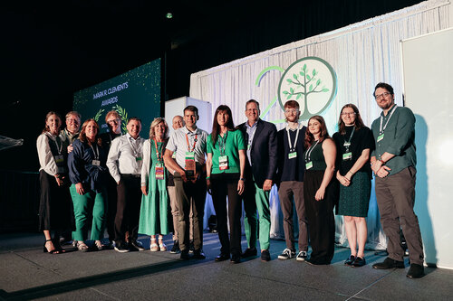 The image depicts a group of people standing together on a stage, likely at an event or awards ceremony. The backdrop features a large, circular logo with a tree design and the number "20," suggesting a 20th anniversary celebration. The text "CHANGE CLIMATE AWARDS" is visible on the backdrop, indicating the event's focus on climate change or environmental achievements.

The individuals are dressed in semi-formal attire, with several wearing green clothing, possibly symbolizing environmental themes. They are standing in a line, smiling, and appear to be posing for a group photo, suggesting a moment of recognition or celebration.

The lighting is bright and focused on the group, highlighting them against the darker background. The atmosphere is positive and celebratory, reflecting a sense of accomplishment and unity.

Overall, the image conveys a professional and environmentally themed event, emphasizing collaboration and recognition in the field of climate change or sustainability.