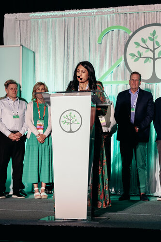 The image depicts a formal event, likely a conference or award ceremony, set on a stage with a podium. A woman stands at the podium, speaking into a microphone. She is wearing a long, patterned dress. Behind her, several individuals are standing in a line, dressed in business or semi-formal attire, including suits and dresses.

The backdrop features a large logo with a stylized tree, suggesting themes of growth, sustainability, or community. The number "20" is integrated into the design, possibly indicating an anniversary or milestone celebration. The predominant colors are green and white, creating a fresh and professional atmosphere.

The lighting is bright, highlighting the speaker and the backdrop, contributing to a formal and celebratory mood. The setting suggests a corporate or organizational event, possibly related to environmental or social initiatives, given the tree symbol and the green color scheme.