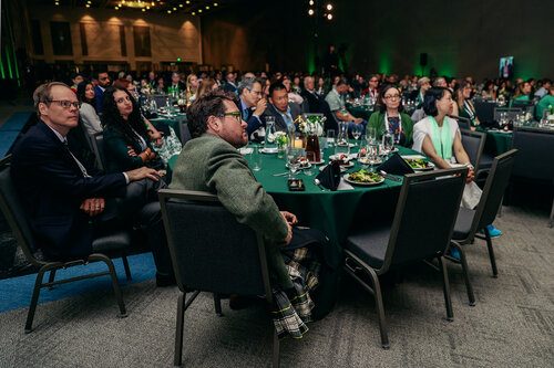 The image depicts a formal event, likely a conference or gala dinner, held in a large, dimly lit banquet hall. The atmosphere is professional and sophisticated, with attendees seated at round tables covered in dark green tablecloths. The tables are elegantly set with wine glasses, plates, and floral centerpieces, suggesting a catered meal is being served.

The attendees, dressed in business or semi-formal attire, are engaged and attentive, facing towards a focal point, possibly a stage or speaker, which is not visible in the image. The lighting is subdued, with spotlights illuminating the ceiling and casting a warm glow over the audience, enhancing the formal ambiance.

The setting is spacious, with rows of tables extending into the background, indicating a large gathering. The walls are adorned with subtle decorative elements, and the overall color scheme is a mix of greens and earth tones, contributing to a calm and professional environment.

The image conveys a sense of networking and professional engagement, typical of corporate events, seminars, or award ceremonies. The presence of diverse individuals suggests inclusivity and a wide-ranging audience, possibly from various sectors or industries.