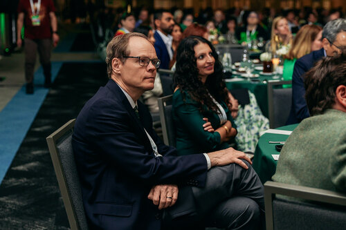 The image depicts a formal event, likely a conference or gala, set in a large indoor venue. The attendees are seated at round tables covered with dark green tablecloths, suggesting a coordinated theme. The lighting is subdued, creating an intimate and focused atmosphere, with spotlights likely illuminating the stage or focal point not visible in the image.

In the foreground, a man in a dark suit and glasses is seated attentively, with his hands resting on his lap, conveying a sense of engagement and interest. Beside him, a woman with long dark hair, wearing a green dress, also appears attentive, with a slight smile suggesting she is enjoying the proceedings. The background shows a diverse group of people, some dressed in business attire, indicating a professional gathering.

The setting includes decorative elements such as floral arrangements and glasses on the tables, adding to the formal ambiance. The presence of lanyards around some attendees' necks suggests this could be a corporate or industry event, possibly involving networking or presentations.

The overall mood is professional and engaged, with a hint of anticipation or interest in the ongoing activities. The event seems to be well-organized, with a focus on networking or knowledge sharing, typical of conferences or formal dinners.