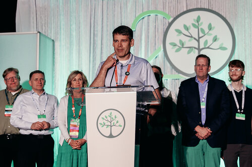 The image depicts a group of individuals standing on a stage, suggesting a formal event or conference setting. The central figure, a man, is speaking at a clear podium with a microphone, indicating he may be delivering a speech or presentation. Behind him, a backdrop features a large circular logo with a stylized tree, possibly symbolizing growth, sustainability, or environmental themes. This logo is also present on the podium, reinforcing its significance to the event.

The individuals are dressed in business or semi-formal attire, with several wearing name badges, suggesting they are participants or organizers of the event. The group appears attentive and engaged, possibly listening to the speaker or preparing to participate.

The color scheme is predominantly green and white, aligning with the tree motif and potentially emphasizing an eco-friendly or nature-related theme. The lighting is bright and focused on the stage, highlighting the speaker and the logo.

The atmosphere conveys professionalism and a sense of occasion, possibly celebrating an anniversary or milestone, as suggested by the number "20" subtly integrated into the backdrop design. This setting could be part of a corporate event, environmental conference, or organizational gathering.