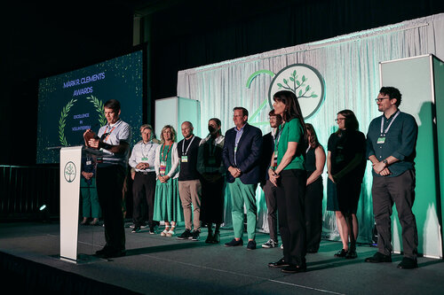 The image depicts a formal award ceremony taking place on a stage. A group of individuals is standing in a line, facing the audience. One person is at a podium, seemingly giving a speech or presentation. The podium features a logo with a stylized tree, suggesting an environmental or sustainability theme.

The backdrop includes a large screen displaying the text "MAJOR CLIMBING AWARDS" along with the words "NARRATIVE" and "VISUAL" beneath it, indicating the categories of awards being presented. The screen is adorned with decorative laurel wreaths, enhancing the formal atmosphere.

The individuals on stage are dressed in a mix of business and casual attire, with some wearing name badges, indicating their participation in the event. The lighting is professional, highlighting the stage and creating a focused, celebratory ambiance.

The predominant colors are green and white, reinforcing the environmental theme. The setting suggests a conference or industry event, possibly related to climbing or outdoor activities, given the context of the awards.

Overall, the image conveys a sense of recognition and celebration within a professional or enthusiast community, with a clear focus on achievements in climbing or related narratives and visuals.