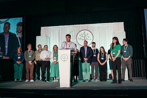 The image depicts a group of individuals standing on a stage, suggesting a formal event or presentation. At the center, a man is speaking at a podium adorned with a circular logo featuring a stylized tree, indicating a possible environmental or organizational theme. The backdrop is composed of vertical white panels with a similar tree logo, reinforcing the thematic focus.

The individuals are dressed in business-casual attire, with a mix of suits, dresses, and slacks, predominantly in shades of green, white, and black, which may symbolize a connection to nature or sustainability. The lighting is bright, highlighting the speaker and the group, creating a professional and focused atmosphere.

The stage setup includes a large screen on the left displaying a live feed of the event, enhancing the visibility of the speaker to a larger audience. The presence of lanyards on the individuals suggests a conference or convention setting.

Overall, the image conveys a sense of unity and purpose, likely related to an environmental or corporate initiative, with an emphasis on collaboration and communication.