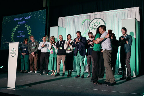 The image depicts an award ceremony on a stage with a group of people standing together. The setting is formal, with a backdrop featuring a large "20" and a tree symbol, suggesting a 20th anniversary celebration. The predominant colors are green and white, creating a fresh and celebratory atmosphere.

On the left side of the stage, a screen displays the text "MAJOR CLEMENTS AWARDS" along with the words "RESILIENCE" and "YOU MATTER," framed by laurel leaves, which are commonly associated with achievement and honor.

The individuals on stage are dressed in semi-formal to formal attire, with several wearing lanyards, indicating they might be participants or honorees at the event. Some people are clapping, while others are taking a selfie, capturing the moment of celebration. The lighting is bright, focusing on the stage and enhancing the festive mood.

The event appears to be a recognition ceremony, possibly for achievements related to resilience or contributions to a cause, as suggested by the text on the screen. The overall atmosphere is joyful and congratulatory, with a sense of community and accomplishment.