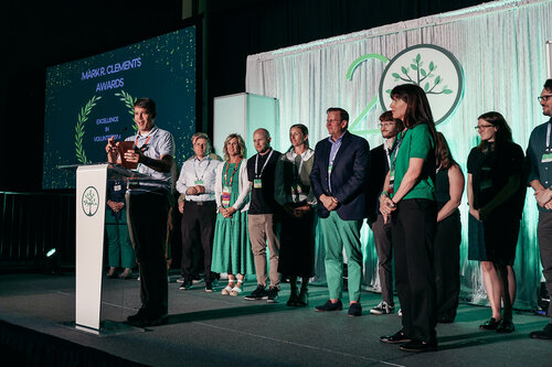 The image depicts a formal award ceremony taking place on a stage. A group of individuals is gathered, with one person standing at a podium, likely delivering a speech or announcement. The backdrop features a large, illuminated "20" with a tree symbol, suggesting a 20th-anniversary celebration. The setting is well-lit, with a mix of green and white lighting that creates a celebratory and professional atmosphere.

The podium displays a circular logo with a tree, matching the backdrop's theme. On a screen to the left, text reads "MIRROR CLEMENTS AWARDS" with "EXCELLENCE" underneath, framed by laurel leaves, indicating the nature of the awards being presented.

The individuals on stage are dressed in formal attire, including suits and dresses, which aligns with the event's formal tone. The audience is not visible, focusing the scene on the presenters and award recipients. The overall mood is one of recognition and celebration, emphasizing achievement and milestone commemoration.