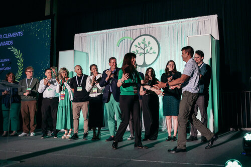 The image depicts a group of people on a stage, likely at an awards ceremony or conference. The setting is indoors, with a backdrop featuring a stylized tree logo and the number "20," suggesting an anniversary or milestone event. The lighting is bright, highlighting the individuals on stage.

In the center, a man and a woman are shaking hands, possibly indicating the presentation of an award or recognition. The woman is dressed in a green top and black pants, while the man is in a light-colored shirt and dark pants. Around them, several people are clapping, suggesting a celebratory atmosphere.

The audience on stage is diverse, with both men and women, dressed in business casual attire. Some individuals are wearing lanyards, indicating they are participants or attendees of the event. The stage design includes green and white colors, which may reflect a theme of growth or sustainability.

To the left, a large screen displays text related to the event, although the specific words are not clear. The overall mood is positive and professional, with a focus on achievement and community.