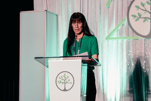 The image depicts a woman standing at a podium, delivering a speech or presentation. She is wearing a green top, and her long hair is down. The podium has a logo featuring a stylized tree within a circle, suggesting themes of nature, growth, or sustainability. Behind her, there is a backdrop with a shimmering curtain and a large numeral "20," possibly indicating an anniversary or special event.

The setting appears to be a formal event or conference, given the professional attire and setup. The lighting is soft, with a greenish hue that complements her outfit and the nature-themed logo, creating a cohesive visual theme. The atmosphere is professional and focused, likely centered around environmental or ecological topics, given the imagery of the tree.

This image might be from a conference, seminar, or anniversary event related to environmental awareness or a similar cause. The overall tone is one of professionalism and dedication to the subject matter being discussed.