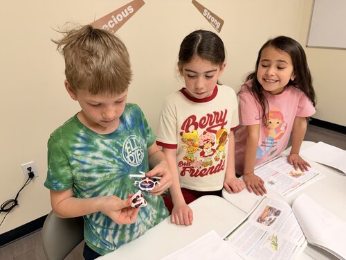 The image depicts three children standing around a table in a classroom setting. The child on the left is wearing a green and blue tie-dye shirt with the text "All Good Friends" printed on it. They are holding a small object, possibly a toy or craft item, and appear focused on it. The middle child is wearing a white shirt with red sleeves and the text "Berry Friends" featuring cartoon strawberries. They are looking at the object held by the first child. The child on the right is wearing a white shirt with a cartoon character and is smiling, looking at the other two children.

The setting includes a table with open books and papers, suggesting a learning or study environment. The walls are cream-colored, and there are motivational words like "precious" and "strong" on the wall behind them. The atmosphere is cheerful and educational, with a sense of camaraderie among the children. The lighting is bright, typical of indoor classroom settings. The scene conveys a theme of friendship and learning, with a playful and engaging mood.