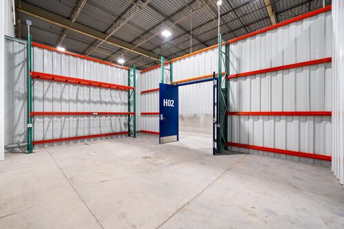 The image depicts an industrial storage unit or warehouse space. The setting is an empty, enclosed area with corrugated metal walls and a high ceiling supported by metal beams. The floor is made of concrete, and the lighting is bright, suggesting artificial overhead lights. 

Prominently, there is a blue door labeled "H102" in white text, which stands out against the otherwise neutral color palette. The door is slightly ajar, adding a sense of depth to the scene. Surrounding the door are metal shelving units with red horizontal beams and green vertical supports, though they are currently empty, indicating the space is either newly constructed or recently cleared.

The atmosphere is utilitarian and functional, typical of a storage or industrial environment. The clean and organized appearance suggests readiness for use or rental. The setting lacks any personal or decorative elements, emphasizing its purpose for storage or industrial use rather than personal or commercial retail.

Overall, the image conveys a sense of spaciousness and potential, with a focus on the structural and functional aspects of the space.
