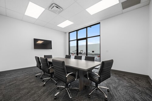 The image depicts a modern conference room with a minimalist design. The room features a large rectangular table at the center, surrounded by eight sleek, black leather office chairs with chrome bases. The chairs are evenly spaced around the table, suggesting a formal meeting setup.

On one wall, there is a large flat-screen television mounted, indicating the room is equipped for presentations or video conferences. The opposite wall has a large window that allows natural light to flood the space, offering a view of an industrial or commercial building exterior, characterized by its metallic facade.

The room's color palette is neutral, dominated by shades of gray and white, contributing to a professional and clean atmosphere. The ceiling is fitted with recessed lighting, providing bright, even illumination throughout the space.

The flooring is covered with a dark, patterned carpet, adding texture and a touch of sophistication to the room. The overall ambiance is one of efficiency and modernity, suitable for corporate meetings or business discussions. The design and furnishings suggest a contemporary office environment, emphasizing functionality and style.