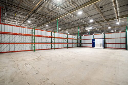 The image depicts a large, empty warehouse interior. The space is expansive, with high ceilings and industrial lighting that provides bright, even illumination throughout the area. The walls are constructed of corrugated metal panels, painted in a light gray color, and are supported by green metal beams. 

There are several red metal shelving units along the walls, but they are currently empty, suggesting that the space is either newly constructed or in transition between uses. The floor is made of concrete, showing some signs of wear and dust, which adds to the industrial feel of the environment.

In the background, there is a blue door, which might lead to another section of the warehouse or an office area. The overall atmosphere is utilitarian and functional, typical of storage or distribution facilities. The setting does not convey any specific cultural or historical context, focusing instead on its practical use as a storage space. The image's clarity and lighting highlight the spaciousness and potential of the warehouse for various industrial or commercial applications.