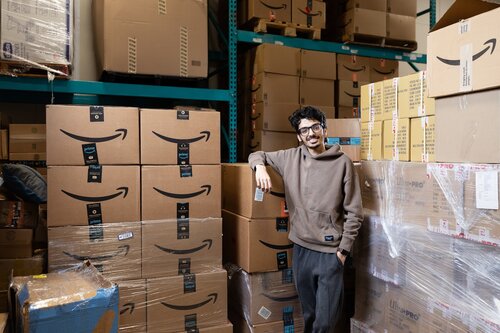 The image depicts a person standing in a storage area filled with numerous cardboard boxes. The boxes prominently feature the Amazon logo, indicating they are likely from the online retail giant. The individual is casually dressed in a brown hoodie and dark pants, leaning against a stack of boxes with a relaxed posture and a smile, suggesting a comfortable and informal atmosphere.

The setting appears to be a warehouse or storage facility, characterized by tall metal shelving units and stacks of boxes reaching up to the ceiling. The lighting is bright, illuminating the space clearly, and the overall color palette is dominated by the brown of the cardboard and the muted tones of the person's clothing.

The image conveys a sense of industriousness and organization, typical of a logistics or e-commerce environment. The presence of Amazon boxes suggests themes of online shopping, distribution, and modern commerce. The atmosphere is one of efficiency and readiness, with the individual possibly representing a worker or someone associated with the handling or management of these goods.

This scene might be associated with themes of retail operations, supply chain management, or the behind-the-scenes workings of a large-scale e-commerce business. The image captures a moment that highlights the scale and reach of contemporary online retailing.