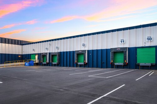 The image depicts a modern industrial loading dock area, likely part of a warehouse or distribution center. The setting features a series of loading bays, each with a green roll-up door, numbered sequentially from 10 to 14. These doors are embedded in a building with a facade of vertical blue and white panels, creating a clean and organized appearance.

The foreground consists of a wide, empty asphalt area marked with white lines, indicating parking or loading zones. The area is devoid of vehicles or people, suggesting either an early morning or late evening time frame when operations are not active.

Above, the sky is painted with a blend of soft pastel colors, including pinks and oranges, typical of a sunrise or sunset, which adds a serene and calm atmosphere to the industrial setting. The lighting is natural, with the colors of the sky reflecting subtly on the building surfaces.

To the left, a blue dumpster is visible near a yellow railing, adding a touch of utility to the scene. The overall mood is one of quiet efficiency, with the well-maintained facilities suggesting a focus on logistics and distribution. The image captures the essence of modern industrial architecture and operations, with a focus on functionality and order.